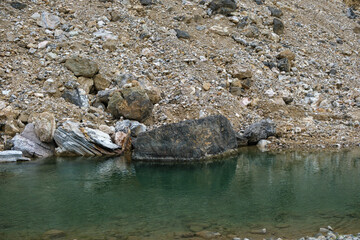 Close-up of a calm, greenish pool of water reflecting a large, dark rock, set against a steep,...