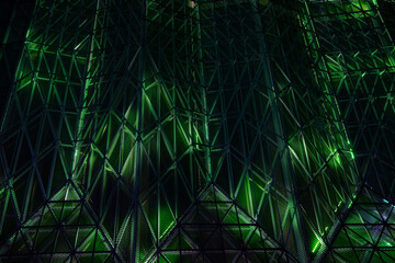 a modern architectural facade at night, featuring a complex lattice of triangular metal frames. The structure is illuminated, creating a geometric pattern of shadows and highlights