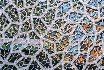 a modern architectural facade featuring a complex, geometric pattern of white, hexagonal and irregularly-shaped frames. The frames enclose various glass panels, creating a mosaic-like effect.