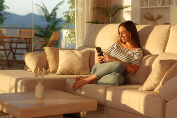 Fototapeta premium Happy woman checking mobile phone at sunset in a living room
