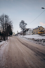 A quiet, snow-covered road winds through a winter landscape in Tromsø, Norway, with traditional houses and bare trees lining the path against a cold, serene sky.