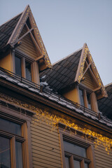 A close-up shot of the roof of a traditional wooden building in Tromsø, Norway, adorned with subtle golden Christmas lights, with snow on the roof and the soft sky above.