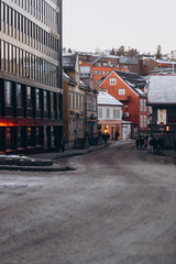 A moody street scene in a city during winter, with slush and snow on the road. A mix of modern and traditional buildings line the street with people walking, capturing a cold urban scene at dusk.
