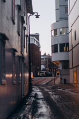 A moody street view in a city during winter, with slush and snow on the road. Buildings and a streetlight frame the narrow perspective, capturing a cold urban scene at dusk.