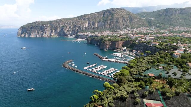 Aerial drone video of the Port of Piano di Sorrento, seen from the sea with the majestic tuff cliffs of the Sorrento coast overlooking the blue waters. A scenic view of the harbor, coastline, and Medi