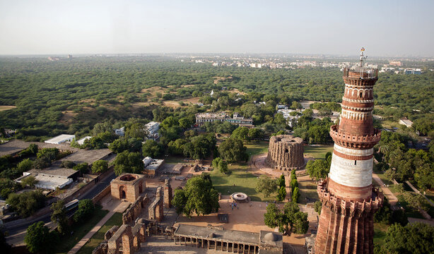 Aerial view of the majestic red sandstone Qutub Minar tower piercing the sky amidst ancient ruins and lush greenery, Delhi, India.