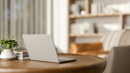 Laptop and books with pot plant on round wooden table and armchair aside bookshelf in living room.