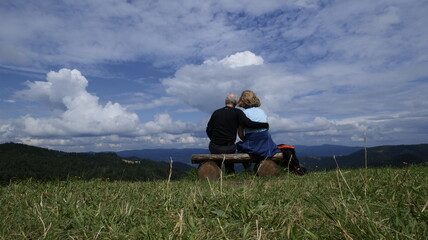 Para osób przytulona do siebie na ławeczce i patrząca na góry w oddali.