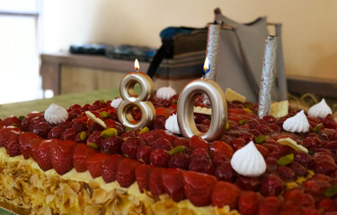 Elegant 80th birthday cake topped with fresh strawberries, raspberries, almonds and meringue kisses, decorated with golden candles shaped as number 80, festive dessert for milestone celebration party