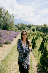 Beautiful young woman wearing sunglasses and casual clothes is walking near lavender flowers and holding smartphone on sunny summer day, enjoying beautiful nature and relaxing atmosphere on sunny day