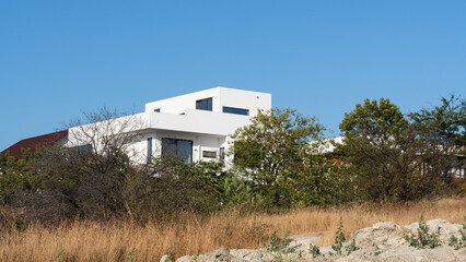 Minimalist modern white house with large windows surrounded by trees and dry grass under clear blue...