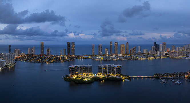 Aerial view of glittering lights of coastal buildings reflecting on the tranquil waters of Dumfoundling Bay, contrasting with the deep blue twilight sky, Aventura, Florida, United States.