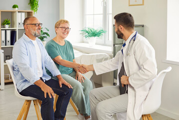 Smiling elderly senior couple meets a doctor in clinic for medical consultation, shaking hands as they discuss care options and treatment. Seniors greeting physician during exam. Medicine concept.