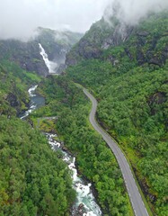 Winding road through a lush mountain valley with a cascading waterfall, captured from a high-angle perspective.