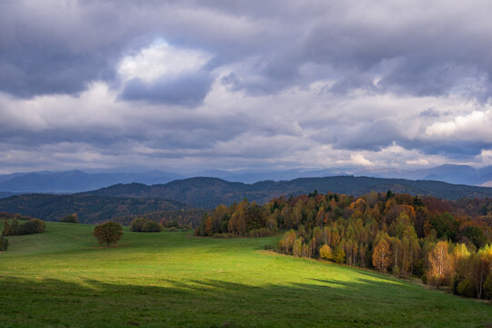 Aerial view of a vibrant green meadow contrasting with autumn-colored trees under a dramatic, cloud-filled sky, Hrochot, Bansk&Atilde;&iexcl; Bystrica Region, Slovakia.