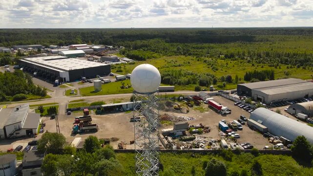 Aerial drone footage orbiting and flying around a modern weather observatory radar dome or white colored radome during a sunny summer day with landscape and city in the horizon at the background.