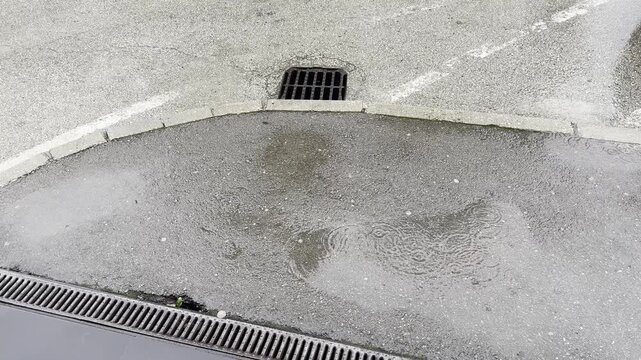 Rainy urban scene with water flowing into drainage grate on wet asphalt road
