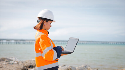 A woman wearing a hard hat and high visibility jacket works on a laptop by the water’s edge,...