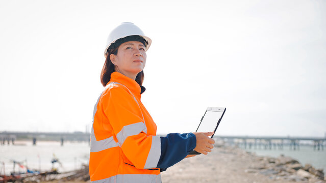 A woman in an orange safety jacket and helmet stands on the shore, using a tablet to oversee activity at a construction site. Bright sunlight enhances the scene, showcasing the work environment