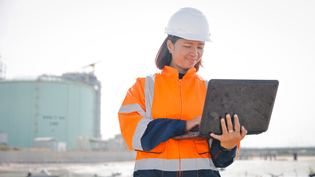 A construction worker, dressed in bright safety gear and a hard hat, is focused on a laptop at a construction site during the day. The background shows industrial structures
