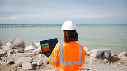 A construction worker in an orange safety vest and white hard hat stands by the water, using a laptop while overseeing activities at a construction site