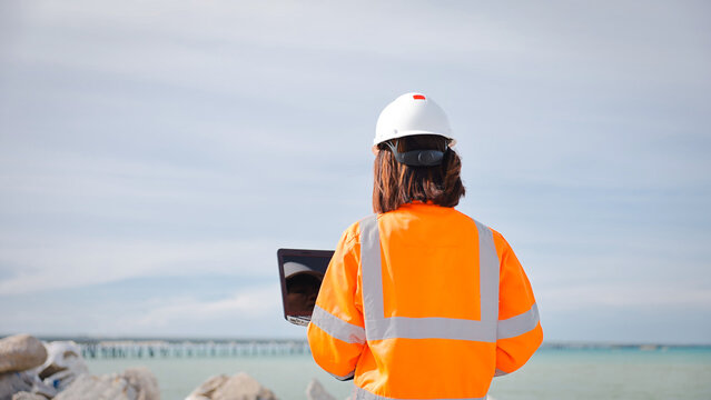 A construction worker in an orange safety jacket and helmet stands at a coastal site. She examines data on her laptop while facing the water, looking for project details