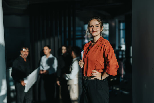 A confident woman in a red blouse stands in a modern office, leading a team meeting. The diverse group of employees collaborates in a professional setting, showcasing teamwork and leadership.