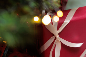 immagine in tema natalizio, con pacco regalo infiocchettato di colore rosso sullo sfondo, con rami di un albero di natale e luci sfuocato in primo piano
