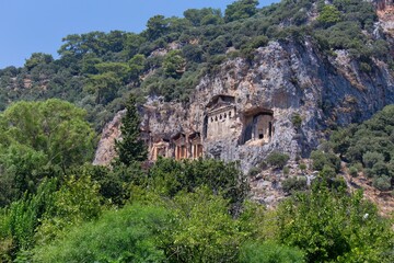Cliffside tombs are carved into the rock face, overlooking a lush, green landscape. The ancient Lycian tombs - Caunos Tombs of the Kings - Dalyan - Mugla - Turkey