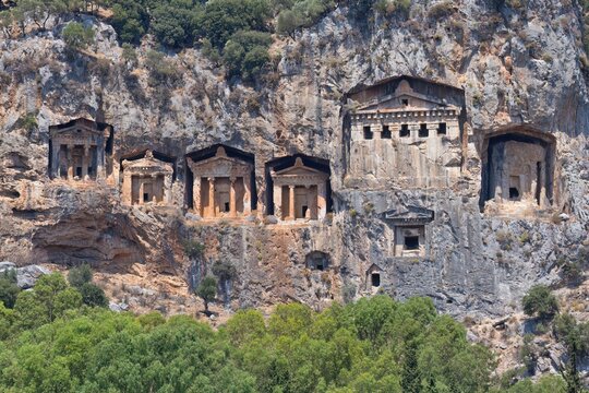 Ancient Lycian rock-cut tombs are carved into a cliff face, featuring temple-like facades. The tombs are set against a rocky backdrop with green vegetation at the base - Caunos Tombs of the Kings 