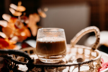 A cozy autumn cappuccino in a glass cup on a rustic wicker tray, surrounded by warm sunlight and seasonal textures. Perfect fall coffee moment.