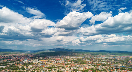 Fototapeta premium An aerial panoramic view of a city surrounded by mountains and greenery under a dramatic cloudy sky.