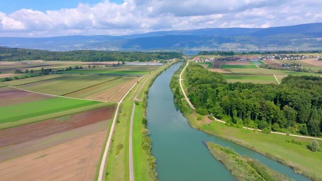 Aerial view of a waterway cutting through vibrant green and brown fields, with a backdrop of distant mountains, Hagneck, Bern, Switzerland.