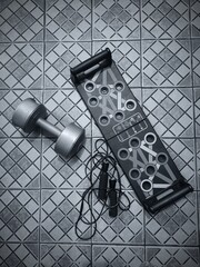Exercise equipment including a dumbbell, jump rope, and push-up board are arranged on a tiled floor.