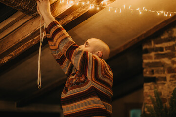 Man in a colorful sweater arranging string lights inside a warmly lit, wooden room.