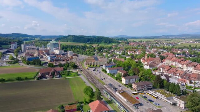 Aerial view of a factory complex with a tall chimney and railway lines contrasting with the bright blue swimming pool, Aarberg, Canton of Bern, Switzerland.