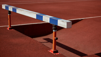 Red track with hurdle casts a distinct shadow under bright sunlight. Concept of hurdle detail, athletics equipment, discipline preparation, sports gear.