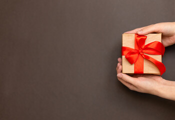 Female hands holding gift with red ribbon in hands, brown background. A beautiful gift decorated with a ribbon and a bow in your hands, space for copy, text and advertising. Banner and background for
