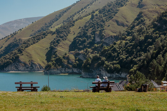 Two people sit on a wooden bench overlooking a serene lake surrounded by green hills in the Caucasus region. The scene is peaceful and picturesque.