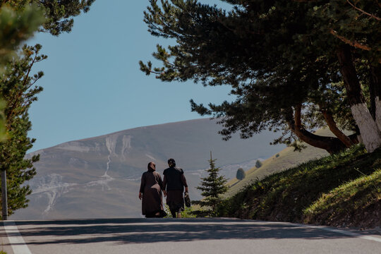 Silhouettes of two people walking along a path in the Caucasus mountains. Pine trees frame the scene with a distant mountain backdrop.