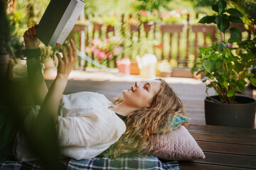 Beautiful young woman reading a book lying on a summer terrace of a country house