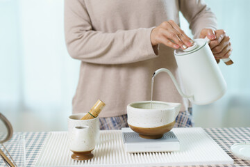 Asian woman preparing traditional Japanese matcha tea at home using bamboo tools, sifting matcha powder with care and precision.