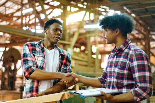 Two African carpenters working together in a sawmill, discussing blueprints, checking wooden pieces, and shaking hands.