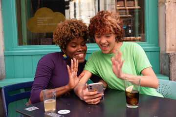 Two friends making a video call and waving at the phone in a bar terrace