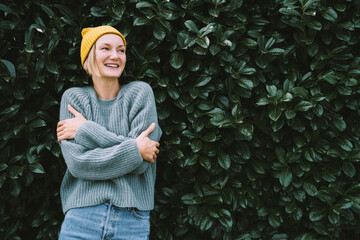 Photo of friendly smiling attractive woman on background of green hedge. Happy confident woman in front of green leaves wall. Thoughtful person outdoors at autumn season.