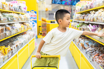 A young boy is seen shopping in a supermarket aisle, reaching for snacks while pushing a small cart.