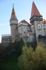 Fototapeta premium A grand medieval castle with tall red-roofed towers, stone walls, and battlements, rising above trees and rocky cliffs, showcasing Gothic architecture and historic charm.