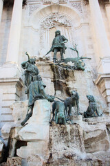 A grand fountain with bronze statues of hunters, dogs, and a deer, set against a carved stone backdrop, symbolizing a royal hunting scene in historic European style
