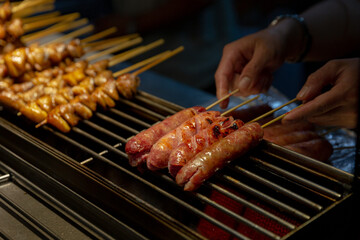 Taiwanese sausages being grilled over open flame at a night market stall, skewers turned by hand as the meat chars and sizzles