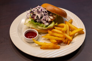 Bagel sandwich served on a white plate with lettuce, purple microgreens, grilled meat patty, creamy sauce, and thick-cut golden fries with ketchup